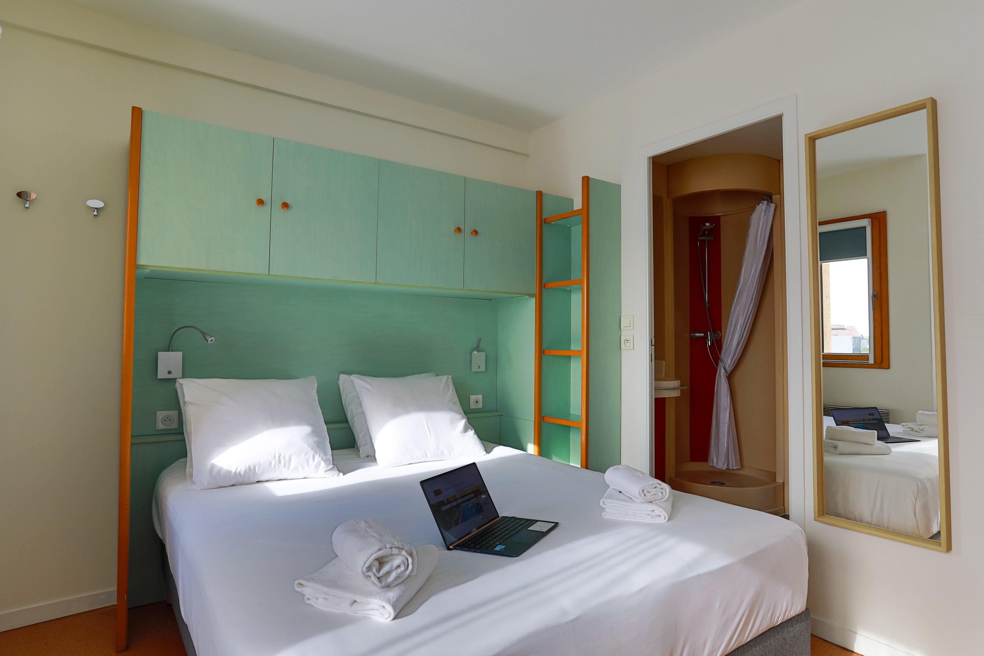 A hotel room with a bed, a laptop, towels, a bathroom with a shower, and a mirror.