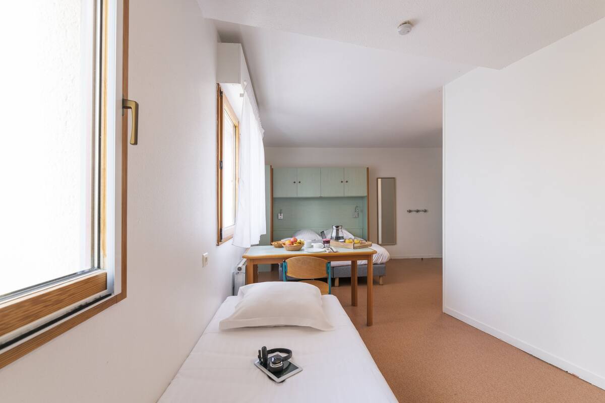 Habitación de hotel moderna con una cama individual, un escritorio con silla, una kitchenette pequeña y un ventanal que proporciona luz natural.