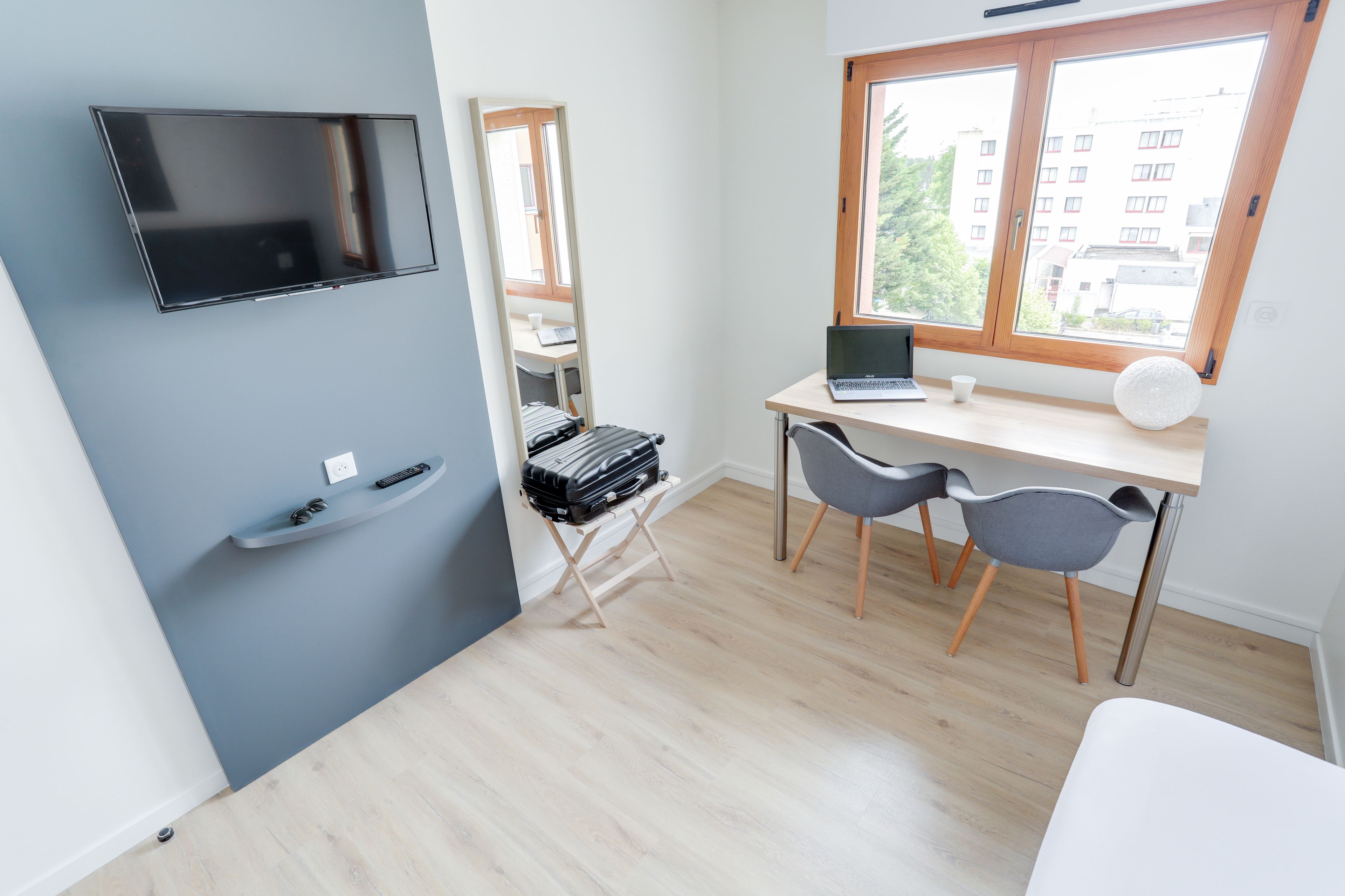 Une chambre moderne équipée d’un téléviseur à écran plat fixé au mur, d’un bureau avec un ordinateur portable, de deux chaises et d’une fenêtre donnant sur des immeubles.