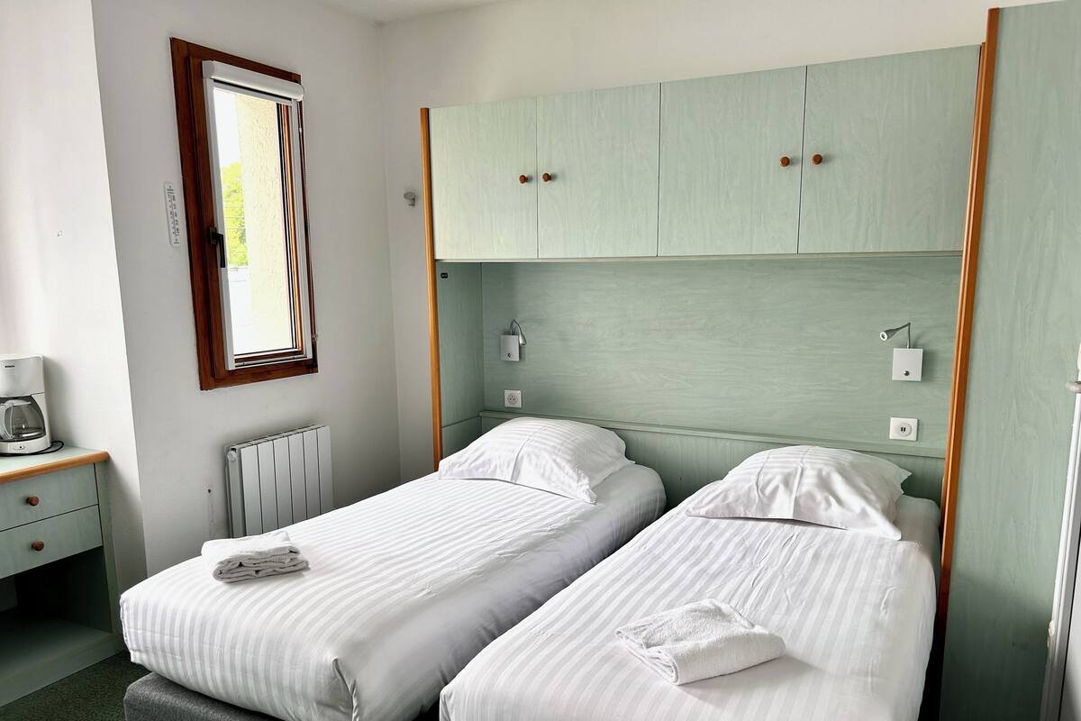 Una habitación con dos camas individuales, una mesita con cafetera y una ventana con persianas.