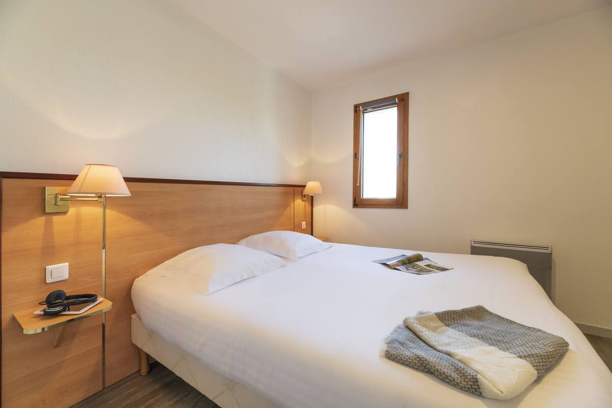 Una cama bien tendida con sábanas blancas, una lámpara de noche y una revista sobre la cama.