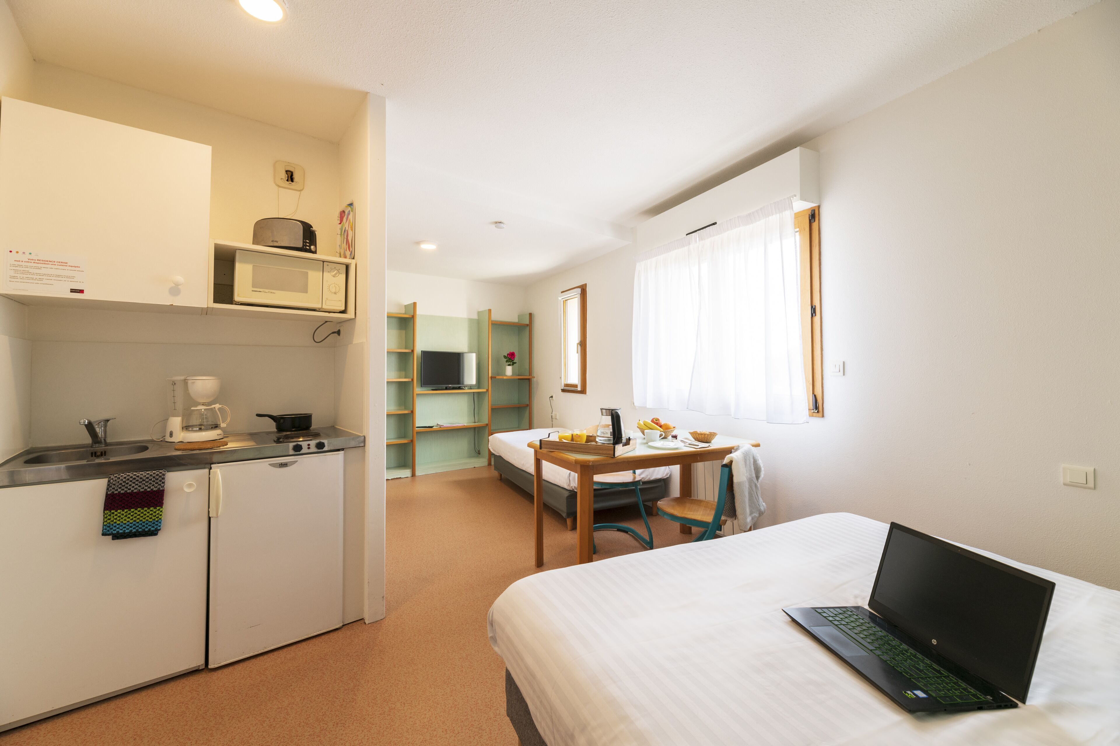 Habitación de hotel compacta con kitchenette, una cama, un escritorio con una computadora portátil y un ventanal con cortinas.
