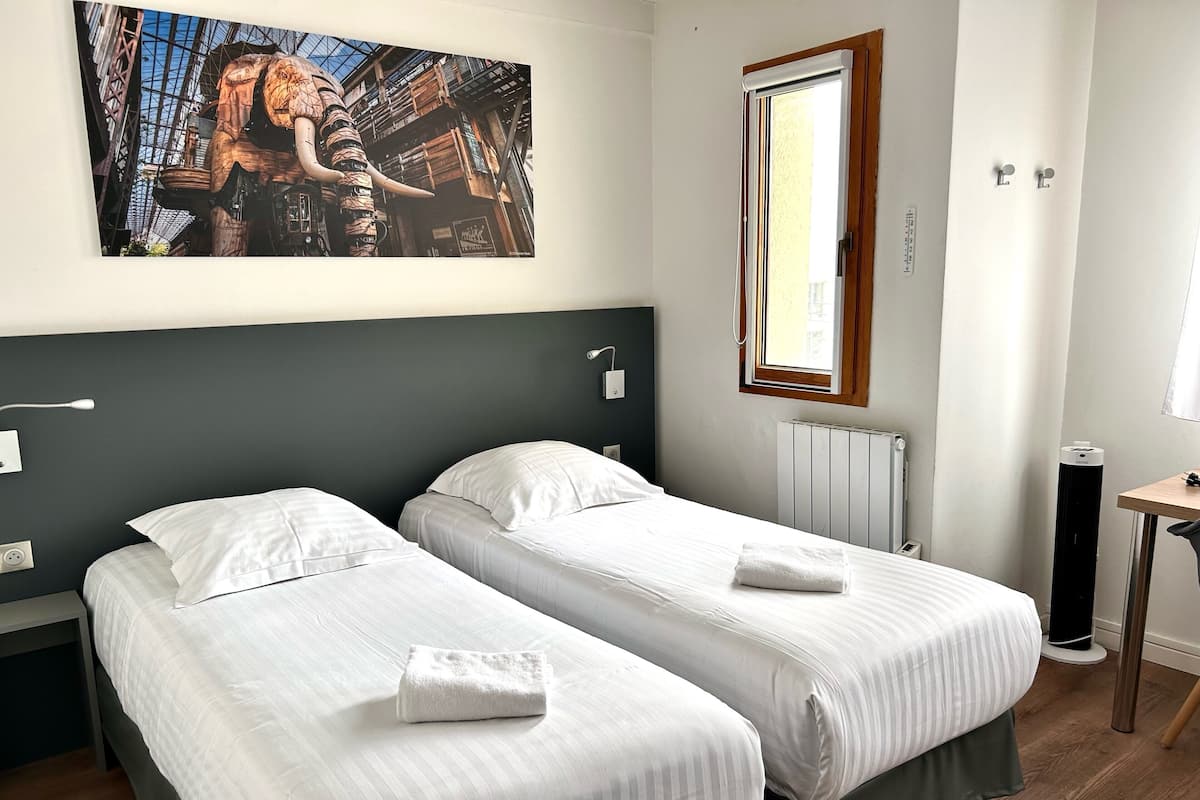 Habitación de hotel con dos camas, un ventanal grande, un radiador y un cuadro en la pared.
