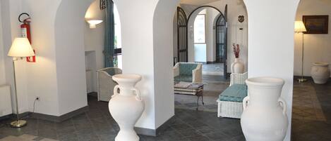 A spacious lobby with white walls, tiled flooring, and large white vases. There are wicker chairs with blue cushions, a floor lamp, and a fire extinguisher.