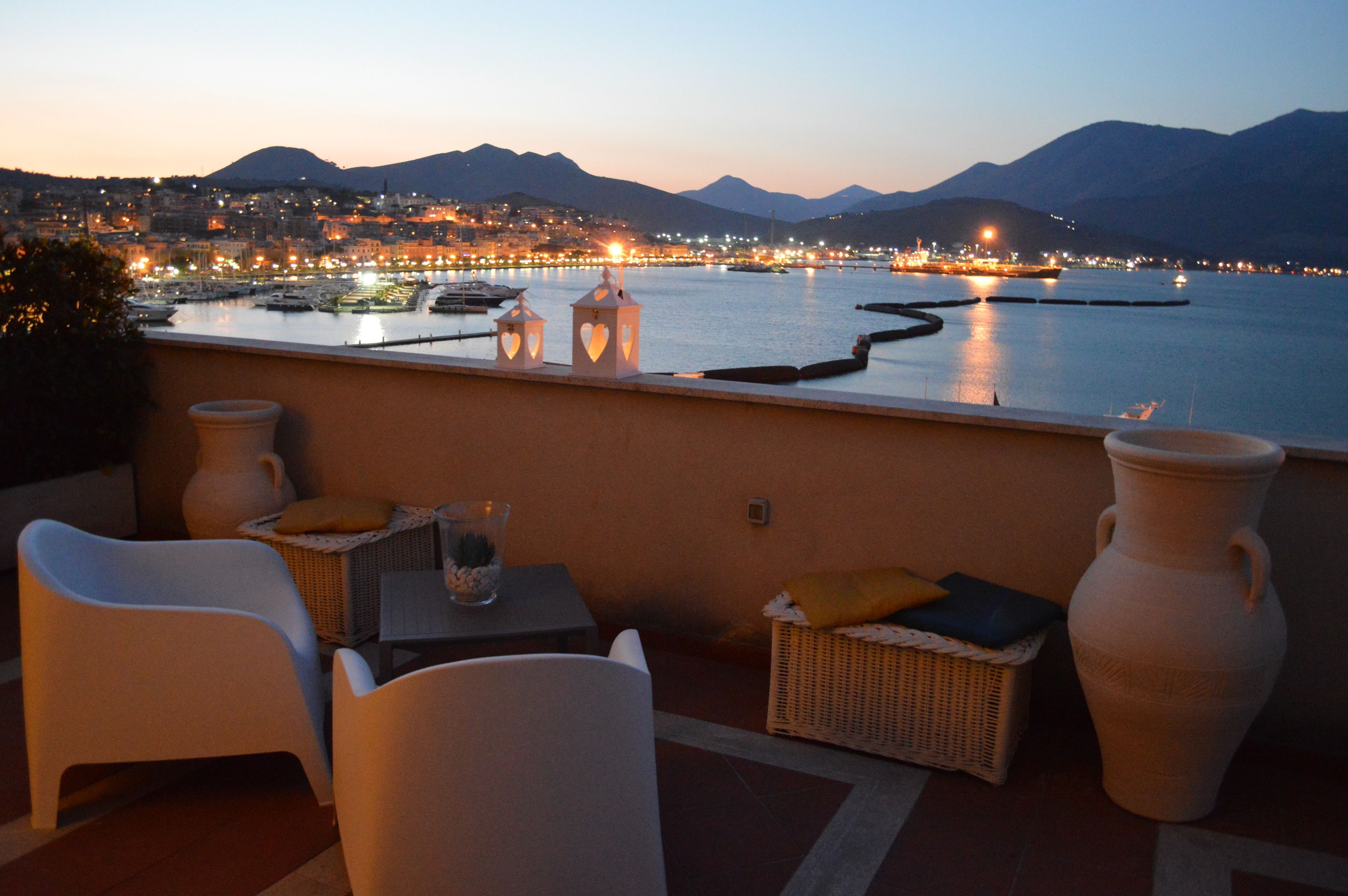 Una terraza con mobiliario de mimbre blanco, dos jarrones grandes y vistas a un paisaje urbano al atardecer.