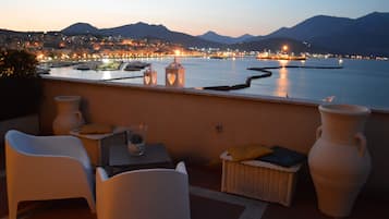 Una terrazza con mobili in vimini bianco, due grandi vasi e vista sulla città al crepuscolo.