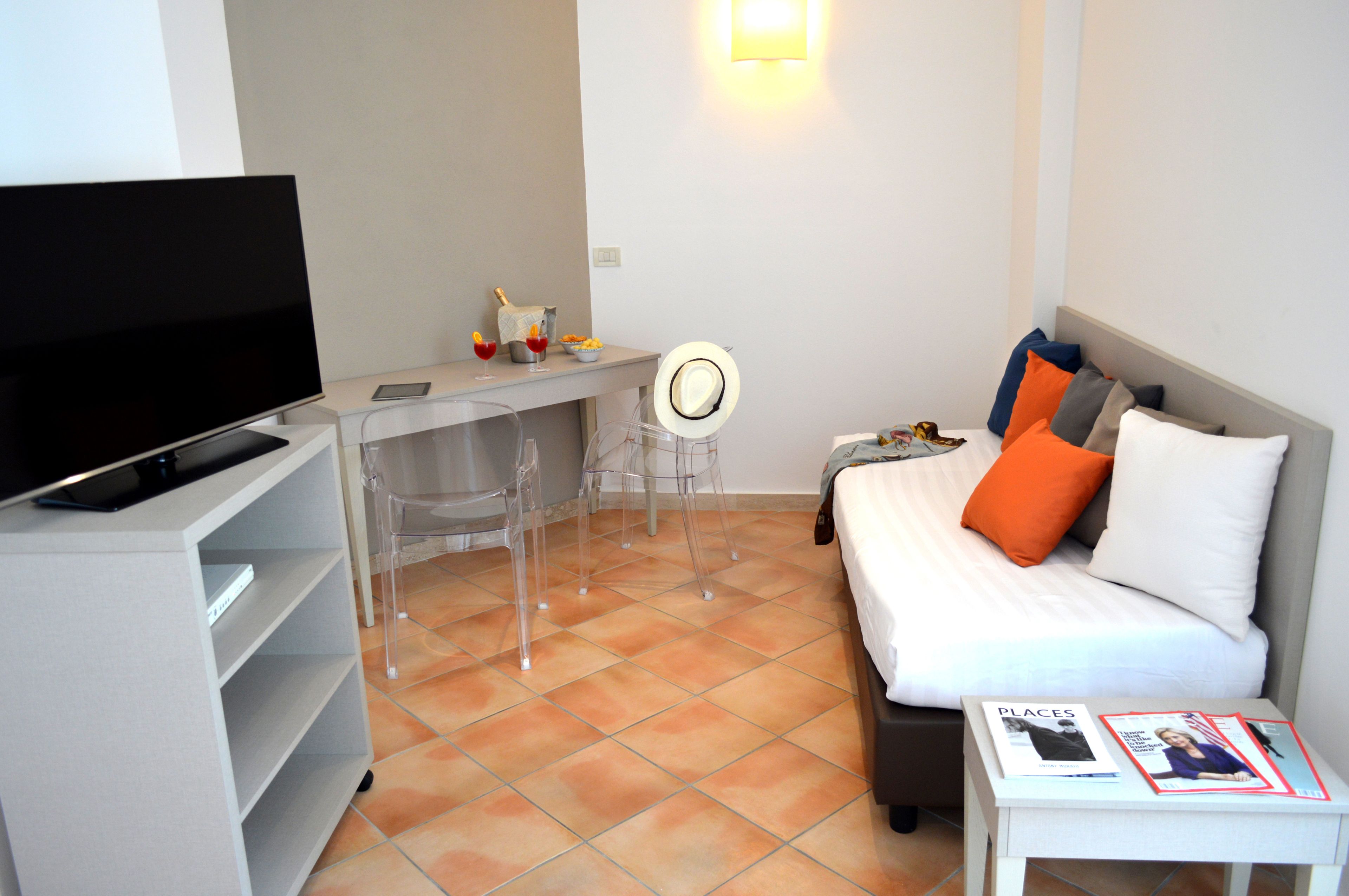 A modern hotel room with a flat-screen TV, a desk with a chair, a bed with orange and grey pillows, and a magazine on the nightstand.