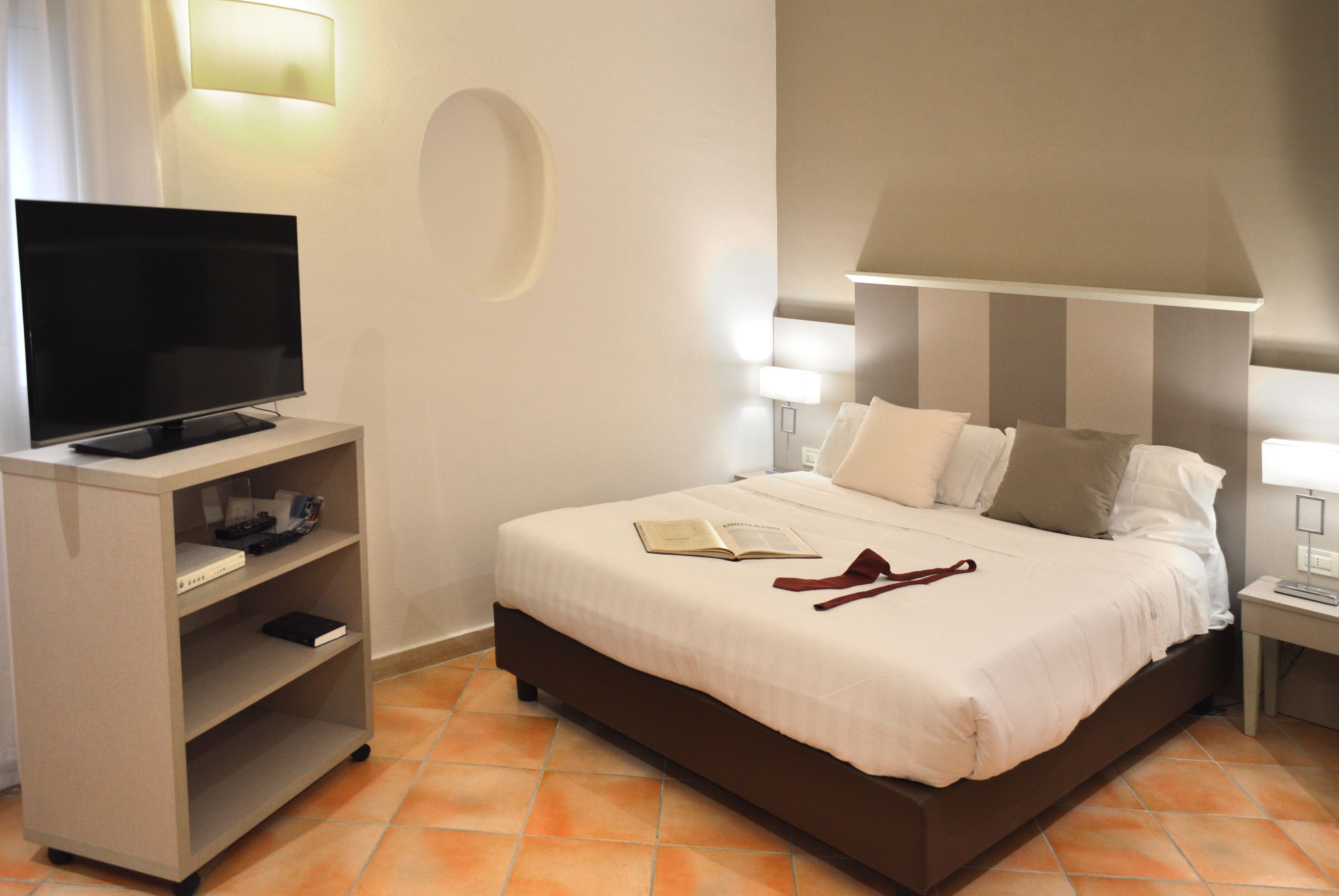 A hotel room with a bed, a TV on a stand, a bedside table, and a lamp.