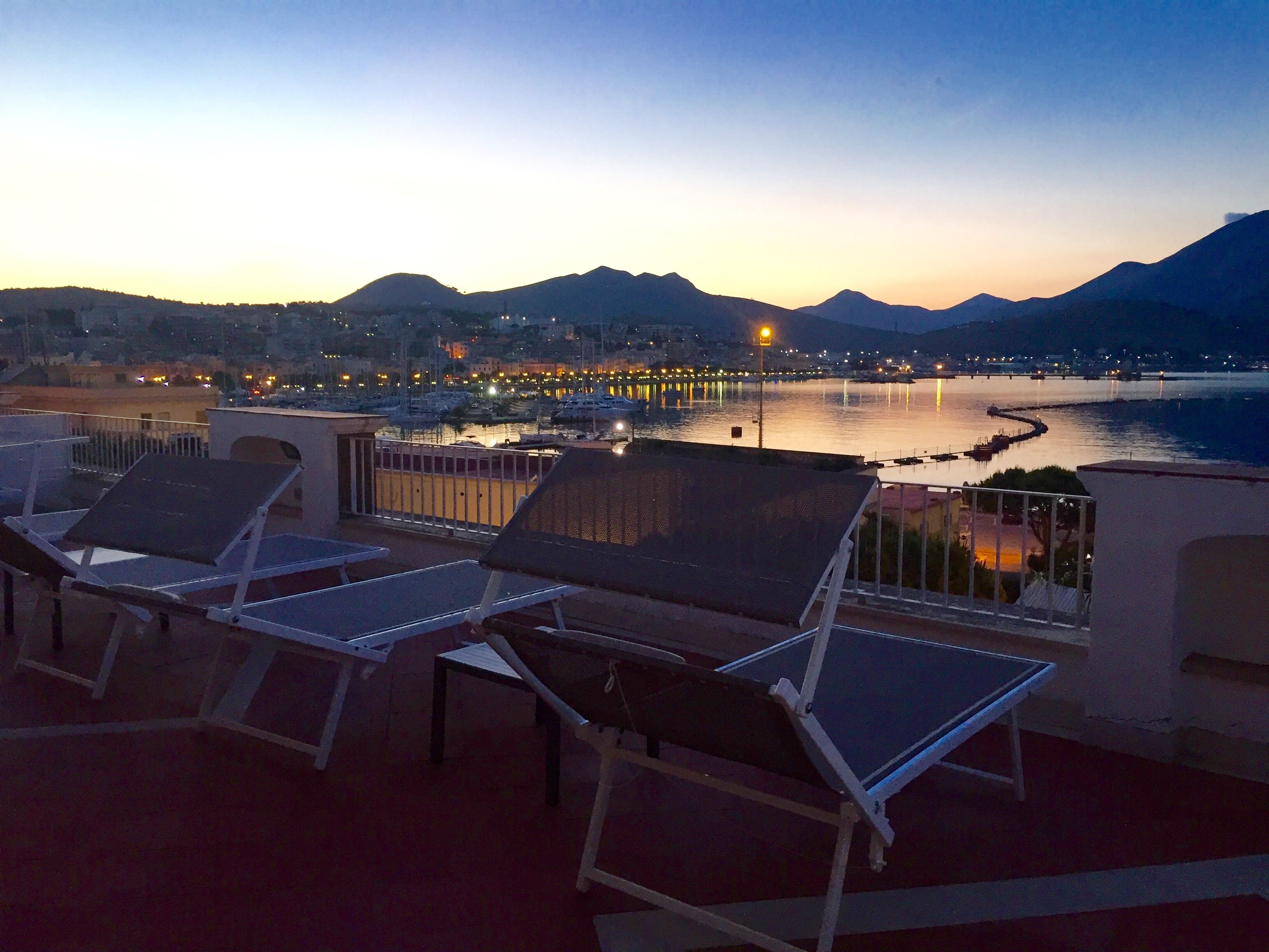 Una terraza con tumbonas que da a la vista de un paisaje urbano frente al mar al atardecer.