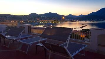 Una terraza con tumbonas que da a la vista de un paisaje urbano frente al mar al atardecer.
