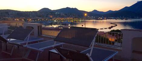 Una terraza con tumbonas que da a la vista de un paisaje urbano frente al mar al atardecer.