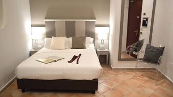 Ein ordentlich bezogenes Bett mit Kissen und einem Buch, ein Paar Schuhe und ein Sessel in einem Hotelzimmer mit Fliesenboden und einer Tür im Hintergrund.