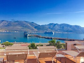 Una terrazza con lettini che si affaccia su un porto con navi e montagne sullo sfondo.