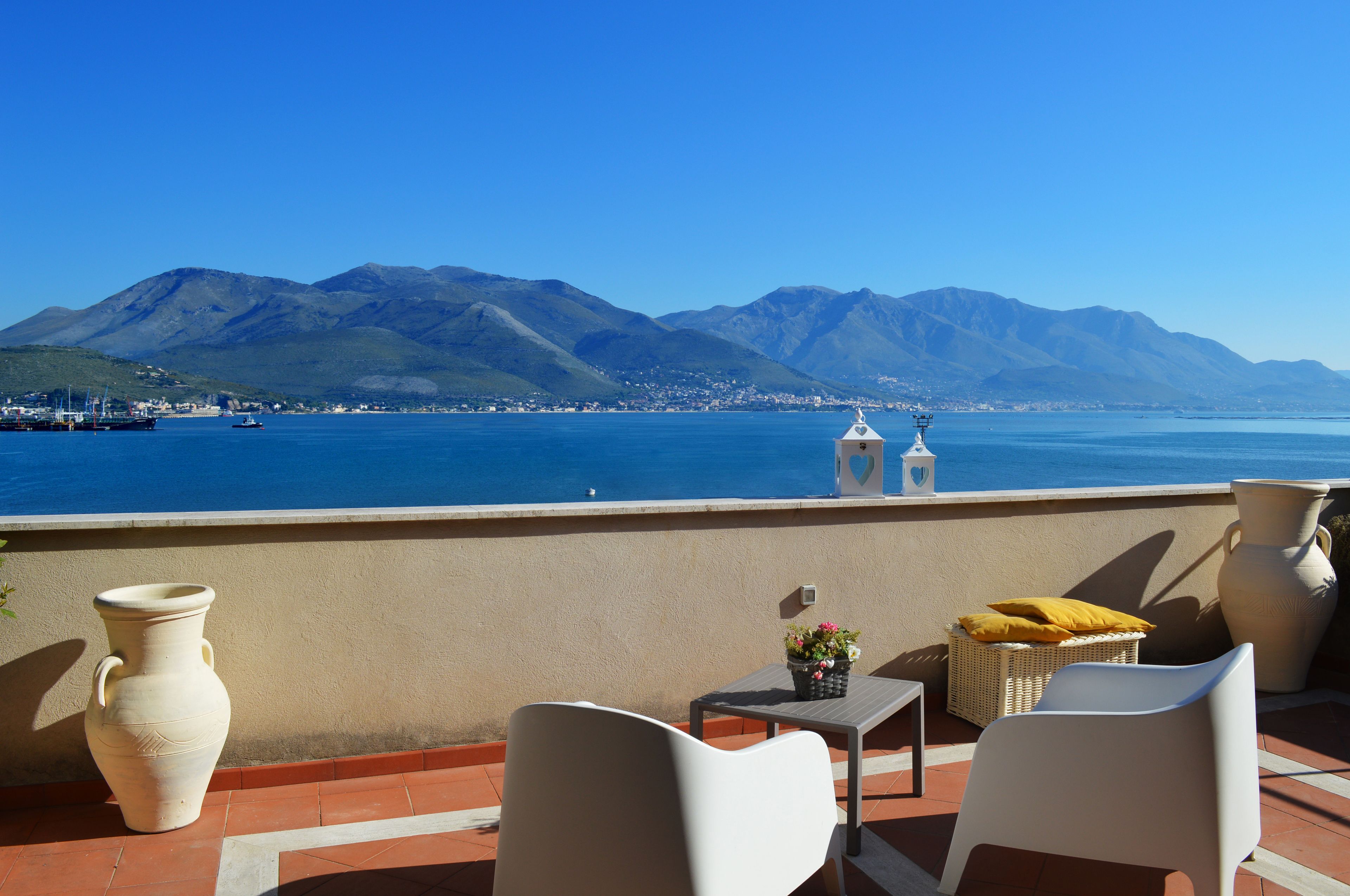 A terrace with a view of the sea and mountains, featuring a white chair, a table with a plant, and a large vase.