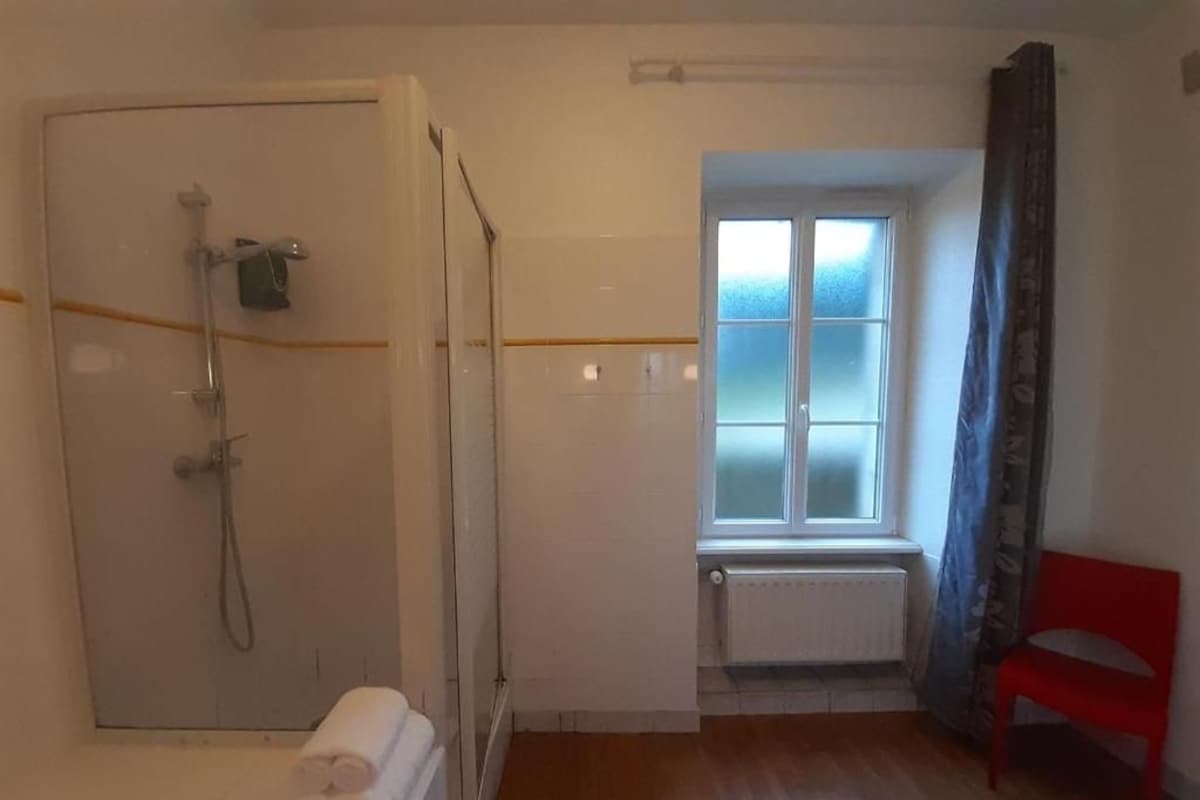 Una habitación pequeña y luminosa con ducha, una ventana con vidrio esmerilado, un radiador y una silla roja.
