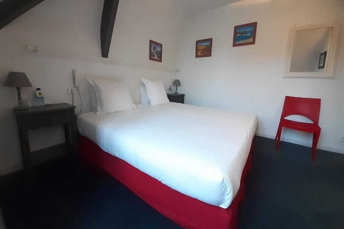 Un dormitorio con cama, mesita de noche, una silla roja y ventana.