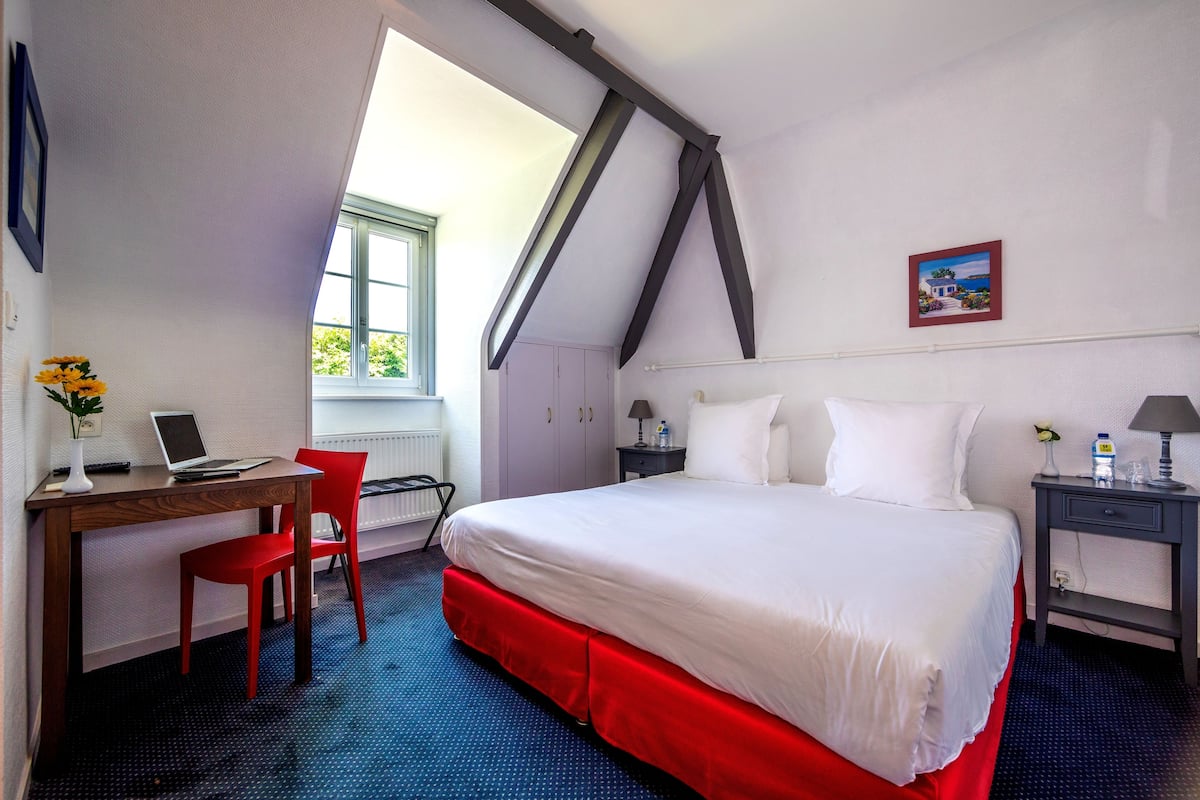 A bedroom with a large bed, a desk with a laptop, a red chair, and a window with a view of greenery.