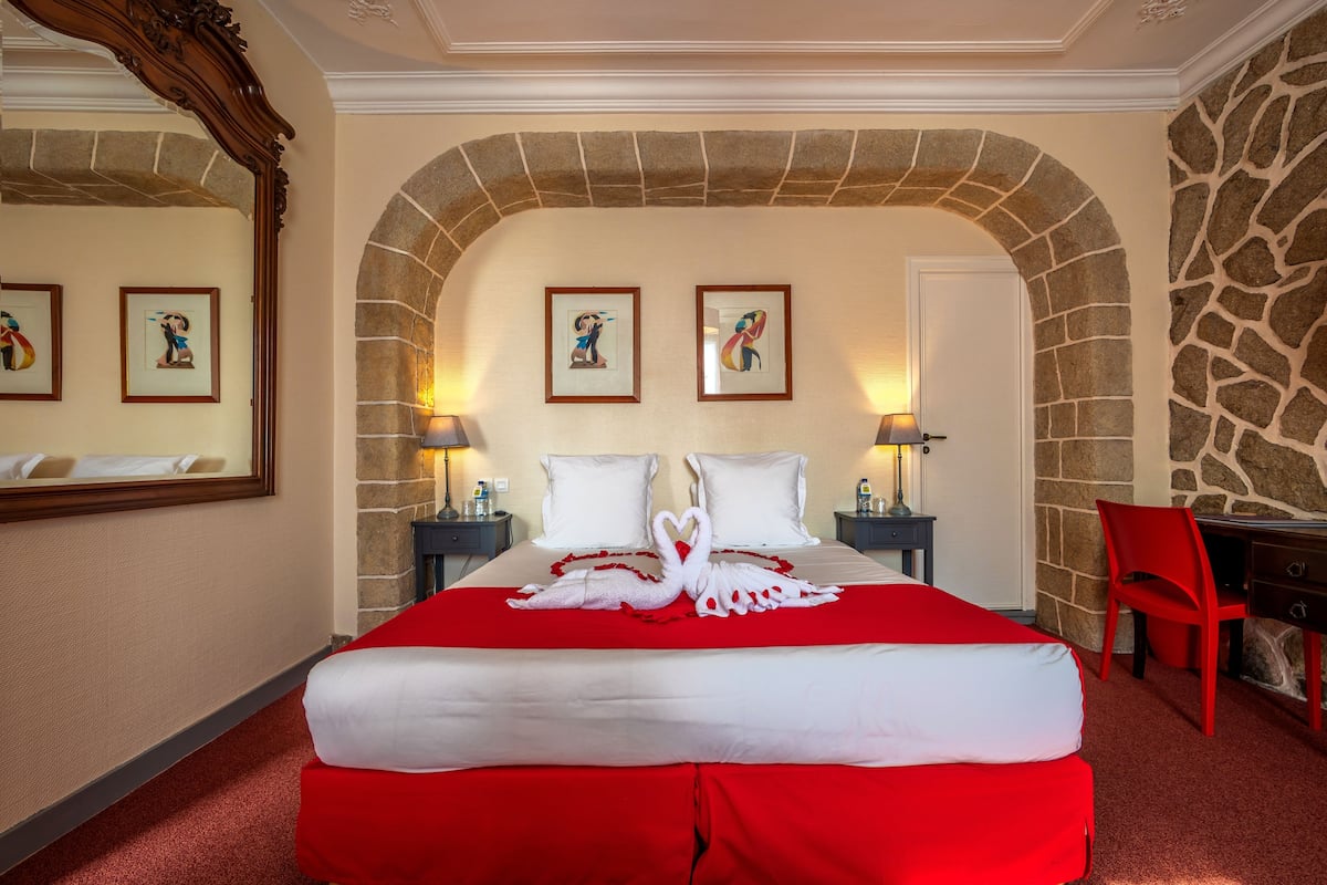 A hotel room with a large bed, a stone archway, two framed pictures on the wall, and a red chair.