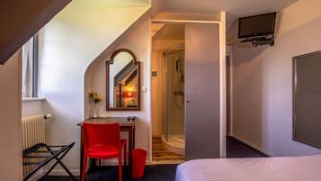 A hotel room with a bed, a desk with a chair, a small table with a vase of flowers, a shower, and a TV mounted on the wall.