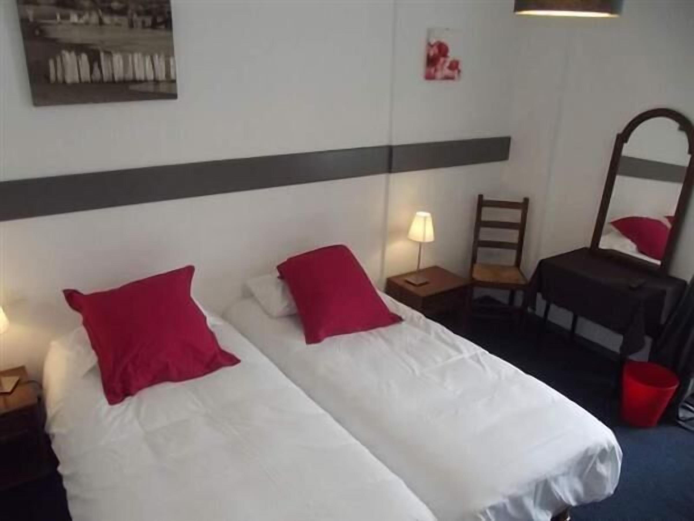 A hotel room with two single beds, each with a red pillow and white bedding. There is a nightstand with a lamp, a chair, and a small table with a mirror.