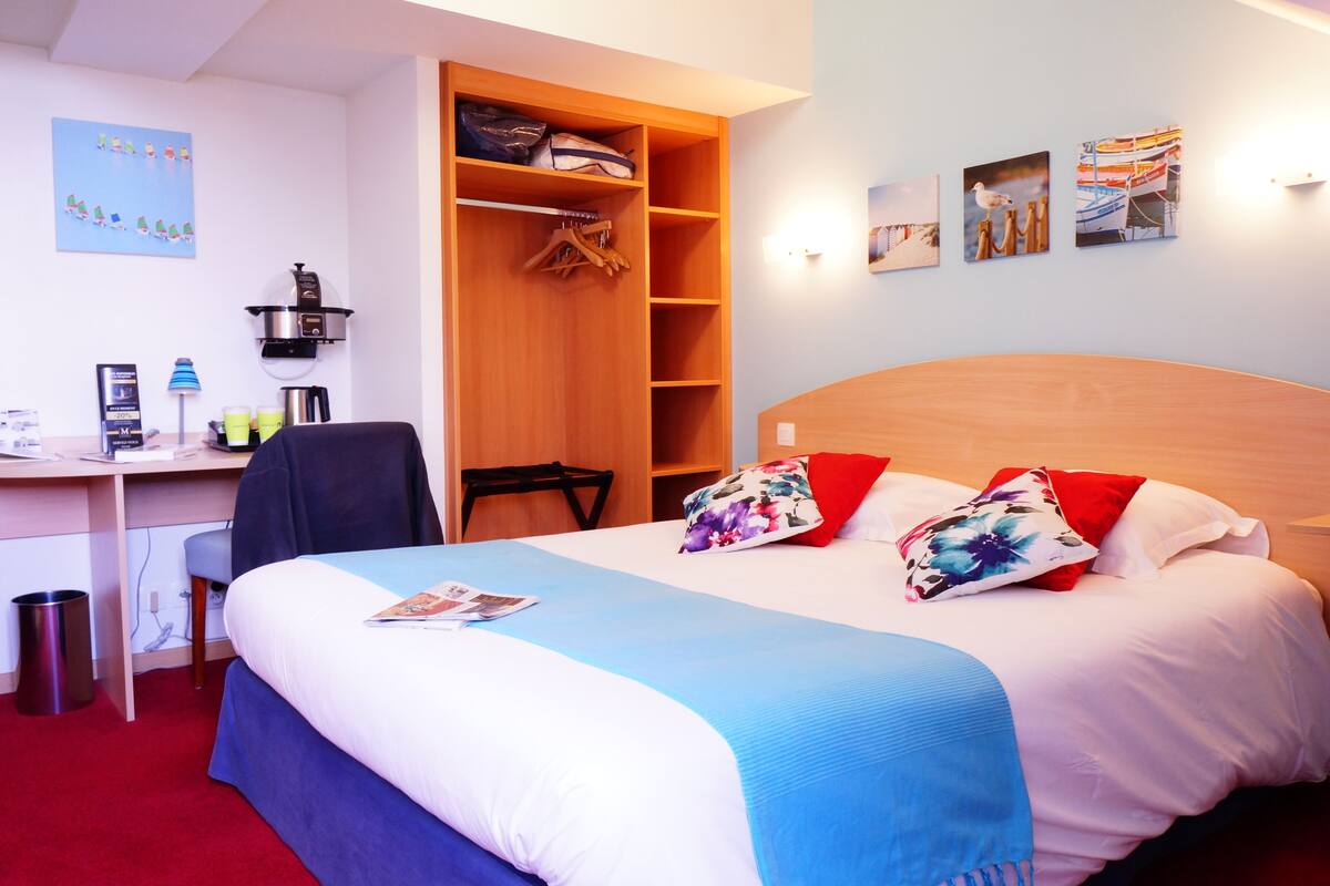 A hotel room with a bed, a desk, a chair, a wardrobe, and a wall with pictures.