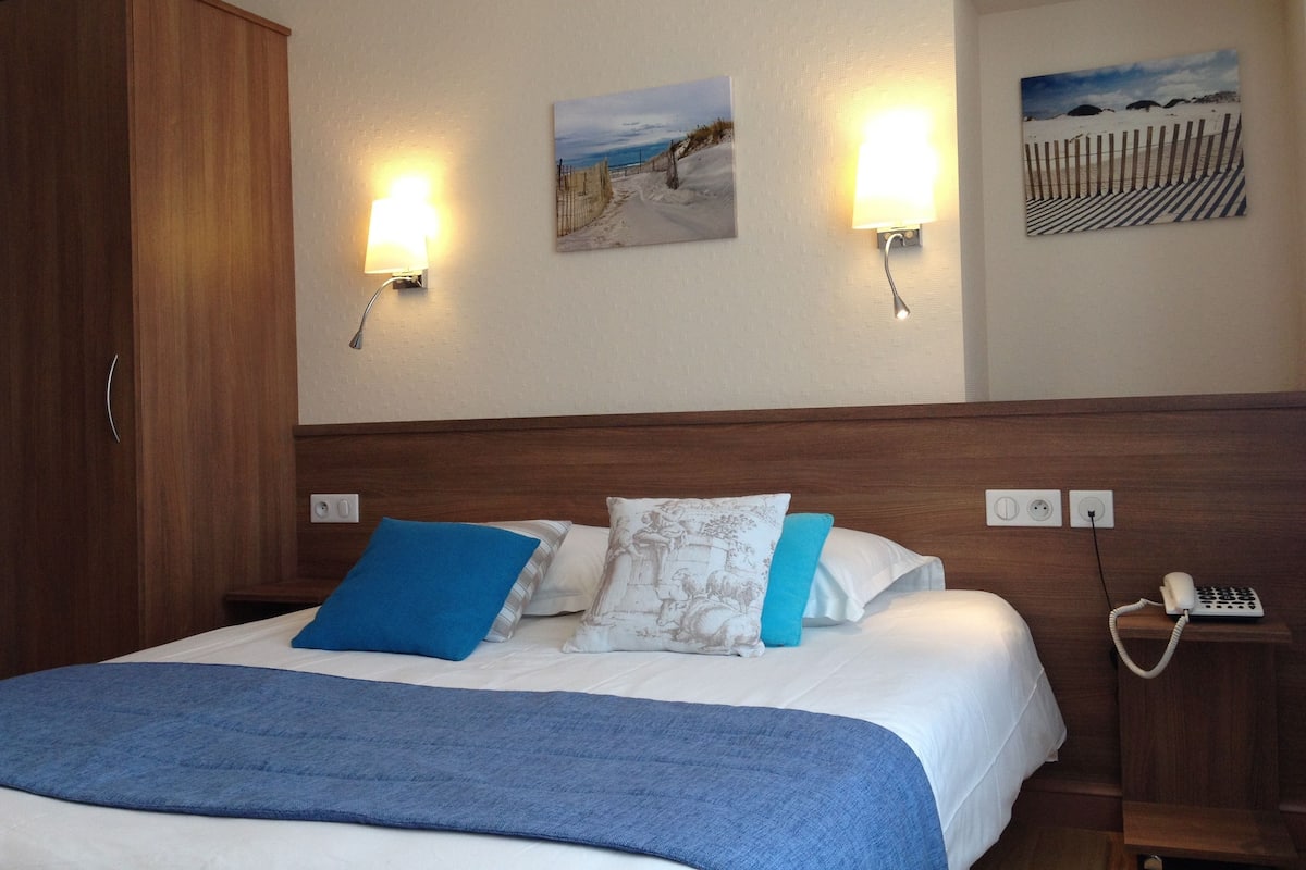 A hotel room with a bed, a wardrobe, two wall-mounted lights, and two paintings.