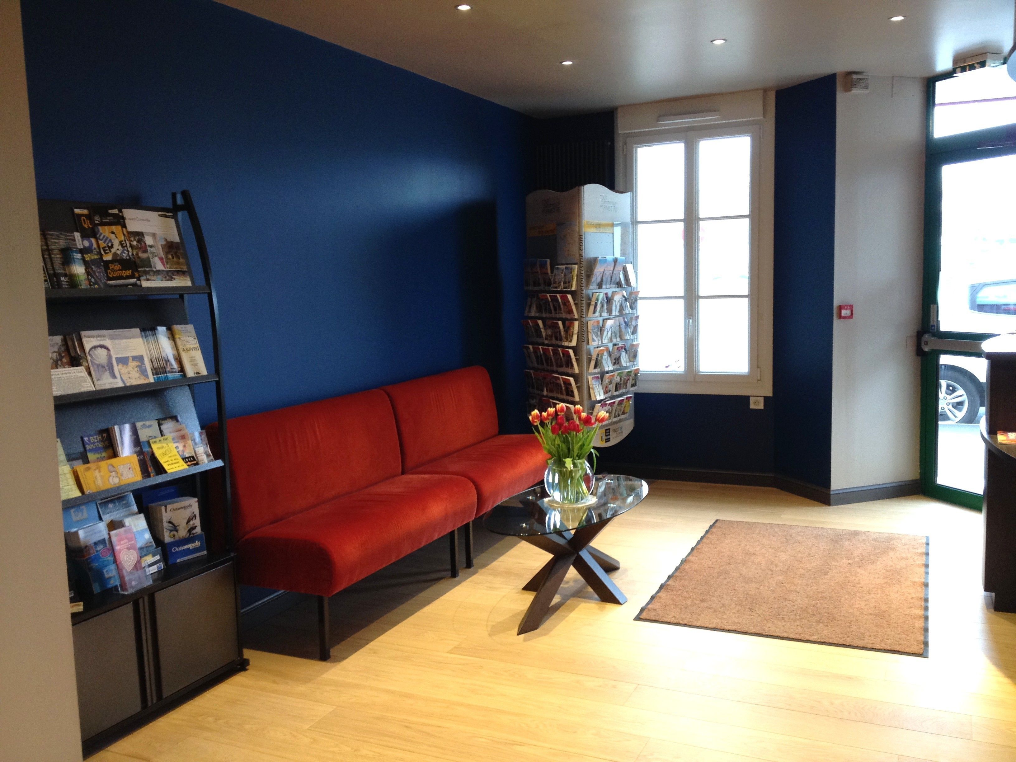 Ein Zimmer mit einer roten Couch, einem Glastisch, einem Bücherregal und einem Fenster mit Blick auf ein Auto draußen.