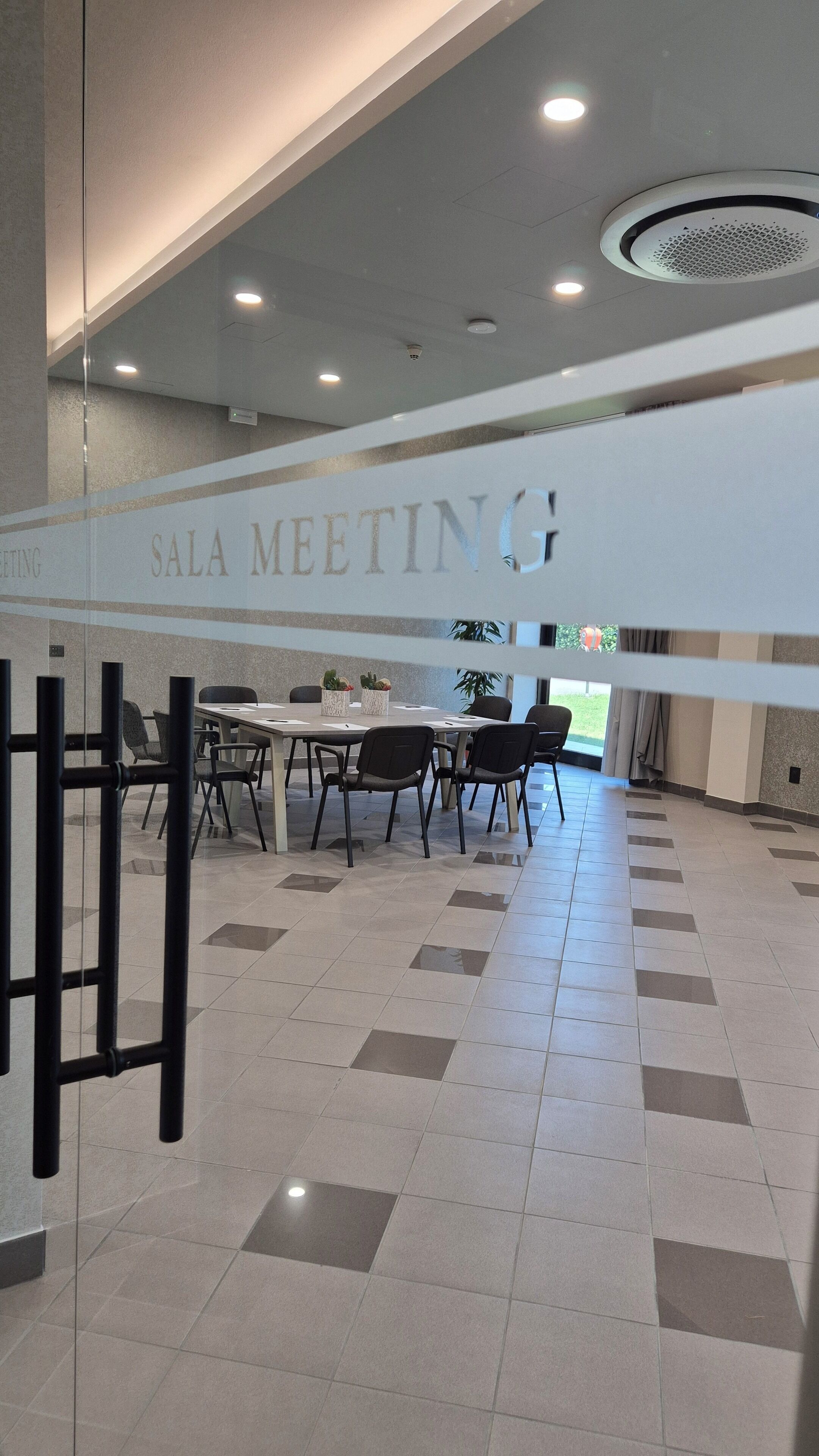 A modern meeting room with a glass partition labeled "SALA MEETING," featuring a table and chairs, recessed ceiling lighting, and a view of the outdoors.