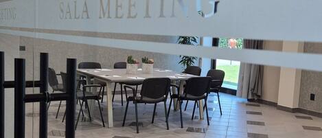 A modern meeting room with a glass partition labeled "SALA MEETING," featuring a table and chairs, recessed ceiling lighting, and a view of the outdoors.