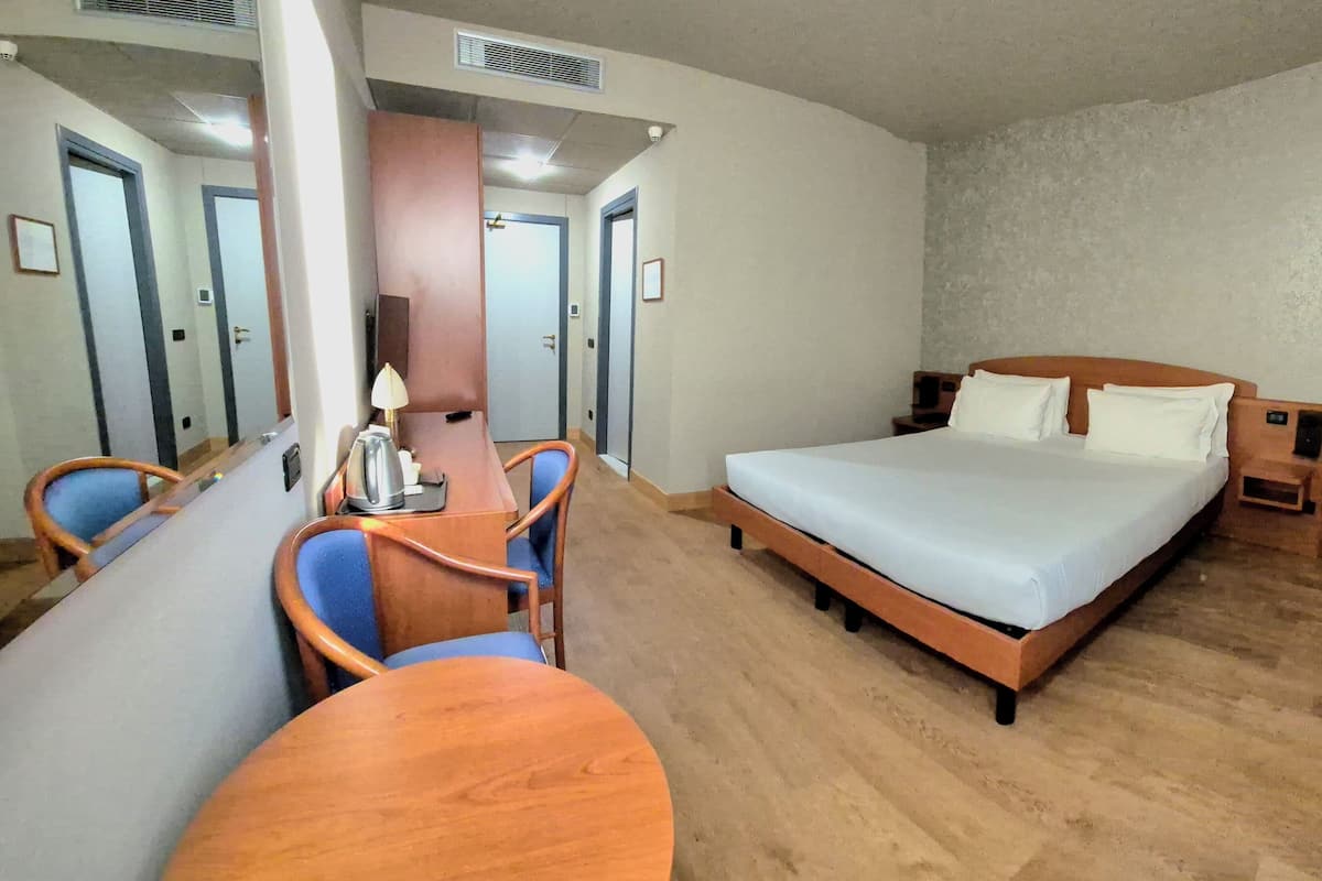 A hotel room with a bed, a desk, two chairs, a lamp, and a mirror.