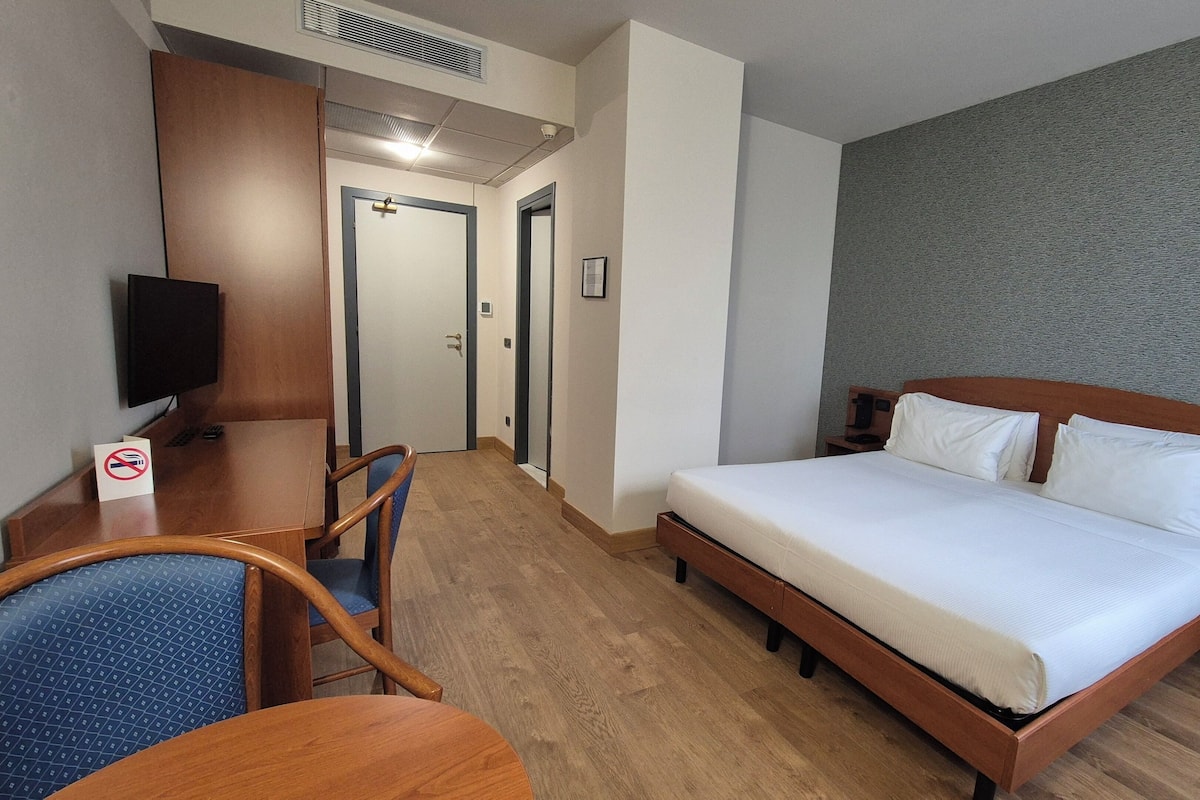 A hotel room with a wooden bed, a desk with a chair, a television, and a no-smoking sign.