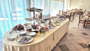 A buffet setup with various food items and utensils on a table, surrounded by chairs and tables in a room with a tiled floor and sheer curtains. - Hotel Le Cerbaie (Altopascio)