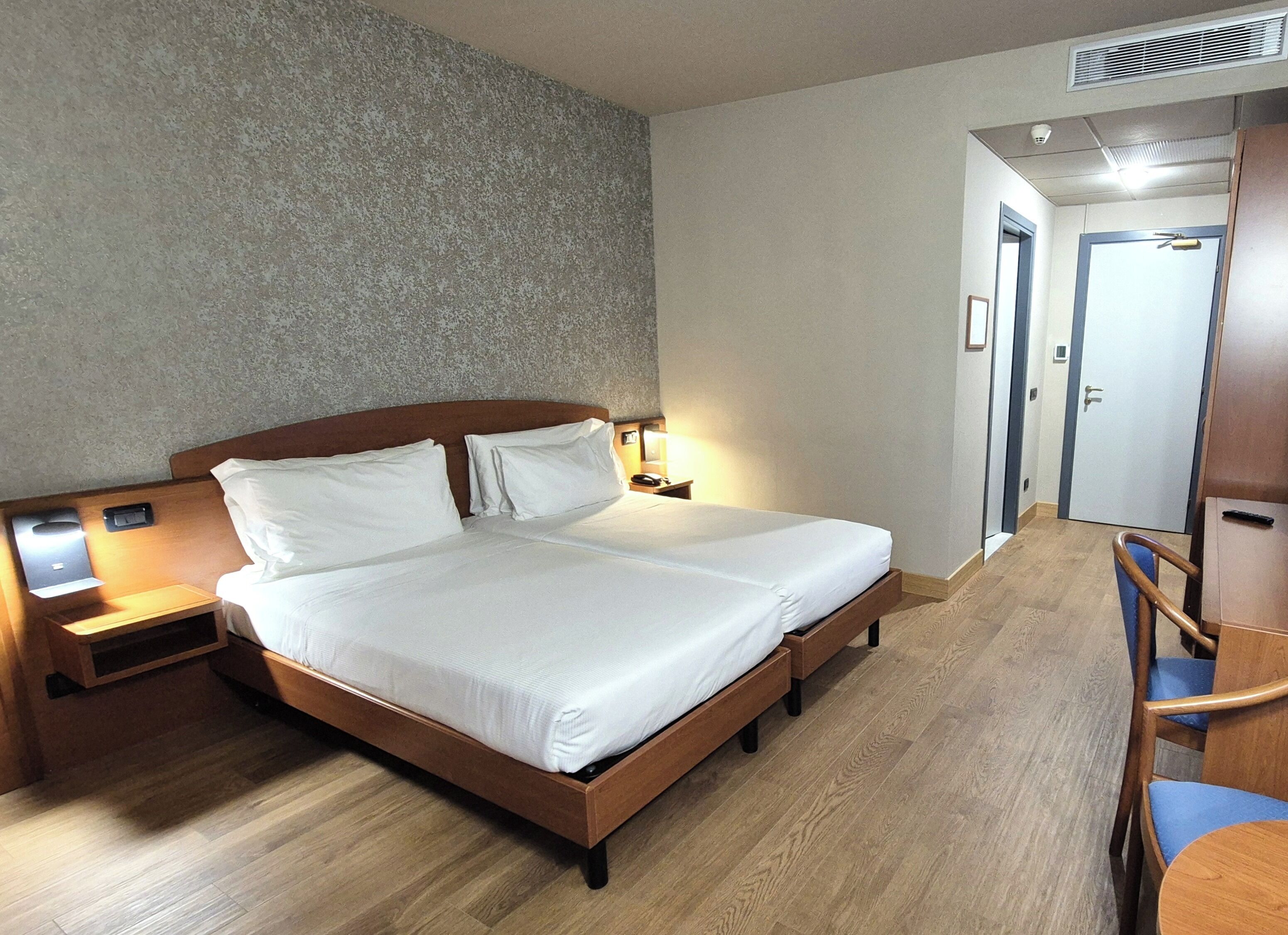 Een hotelkamer met een tweepersoonsbed, een houten hoofdeinde, nachtkastjes met lampen, een houten bureau met een stoel, en een deur die naar een andere kamer leidt.