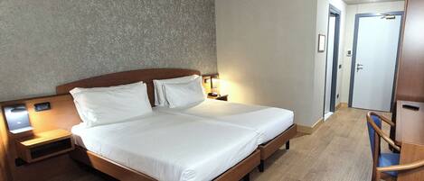 A hotel room with a double bed, wooden headboard, bedside tables with lamps, a wooden desk with a chair, and a door leading to another room.
