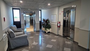 A modern hotel lobby with a reception desk, a seating area with chairs, a potted plant, a fire extinguisher, and an elevator. - Hotel Le Cerbaie (Altopascio)