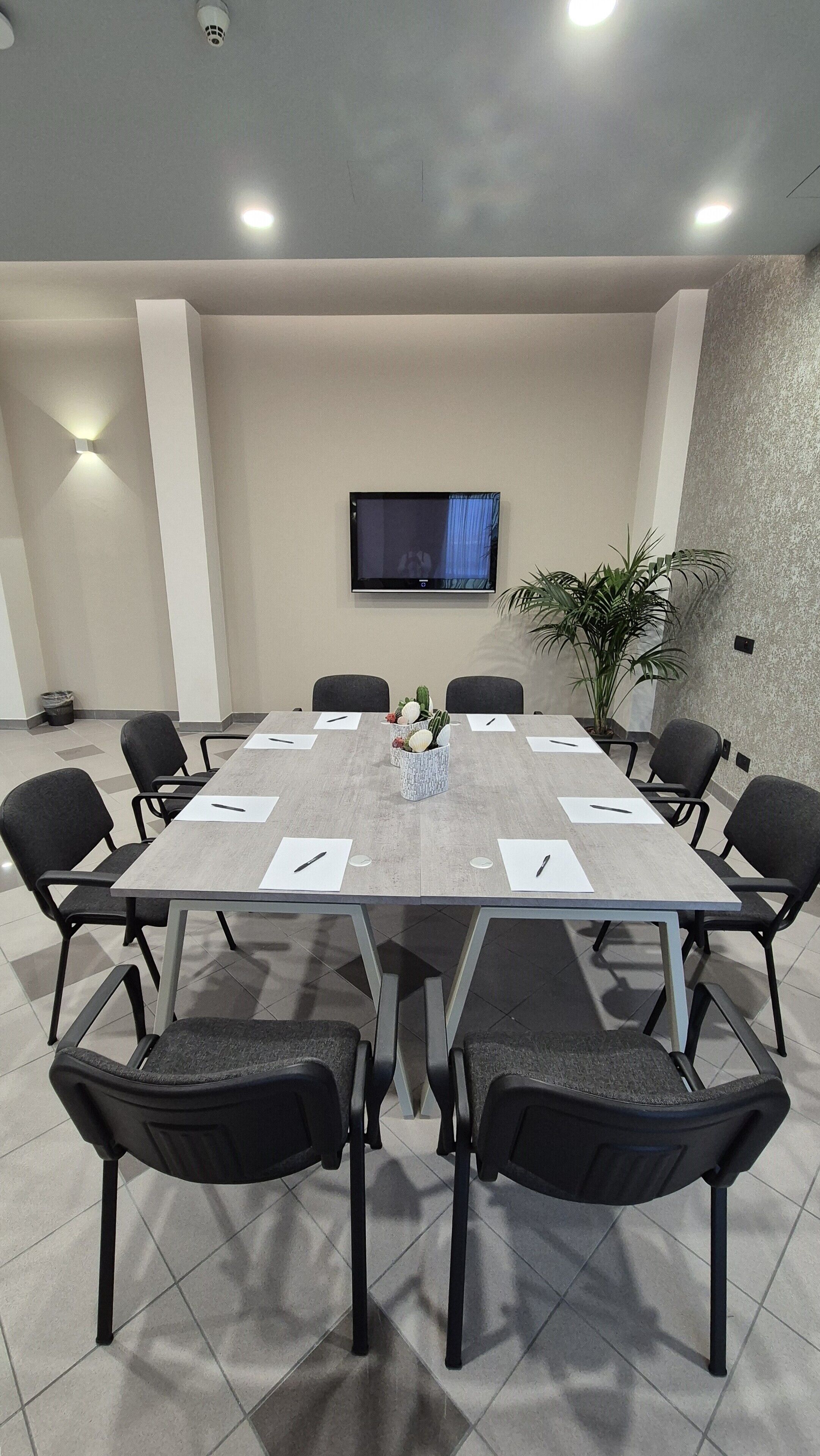 A conference room with a long table, black chairs, a flat-screen TV on the wall, and a potted plant.
