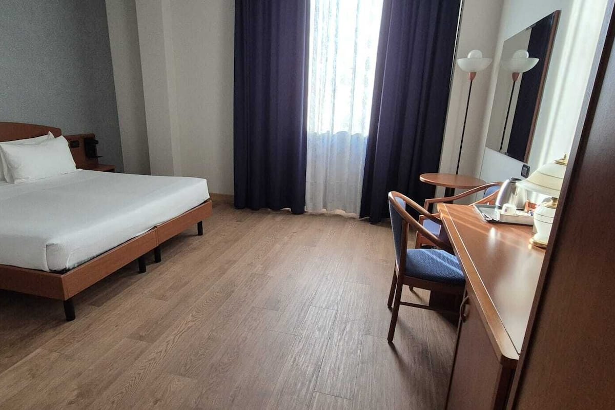 A hotel room with a bed, a desk, a chair, a mirror, and a window with curtains.