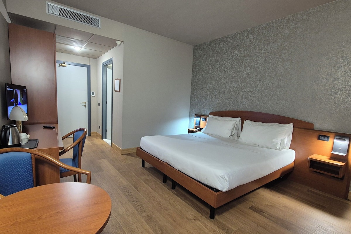 A hotel room with a bed, bedside table, lamp, chair, desk, television, and a door leading to another room.