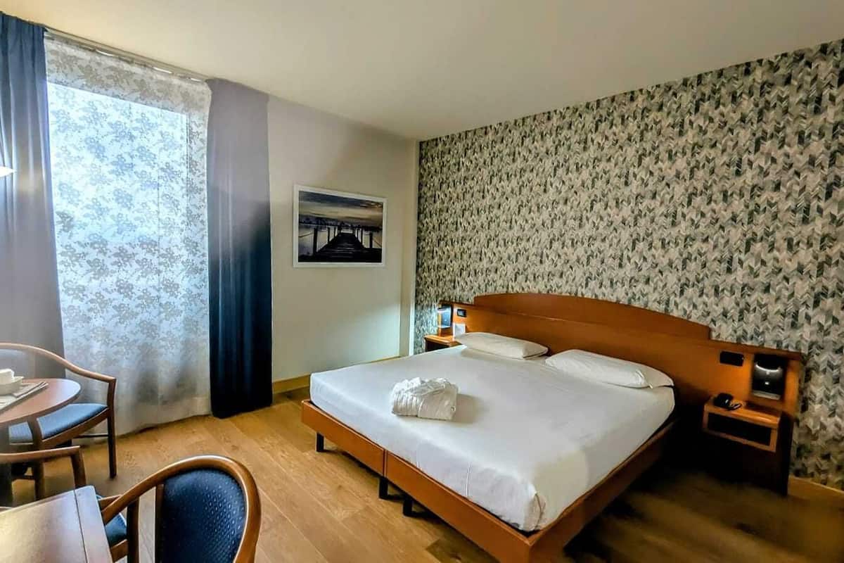 A hotel room with a bed, bedside table, lamp, chair, and a view of the outdoors.