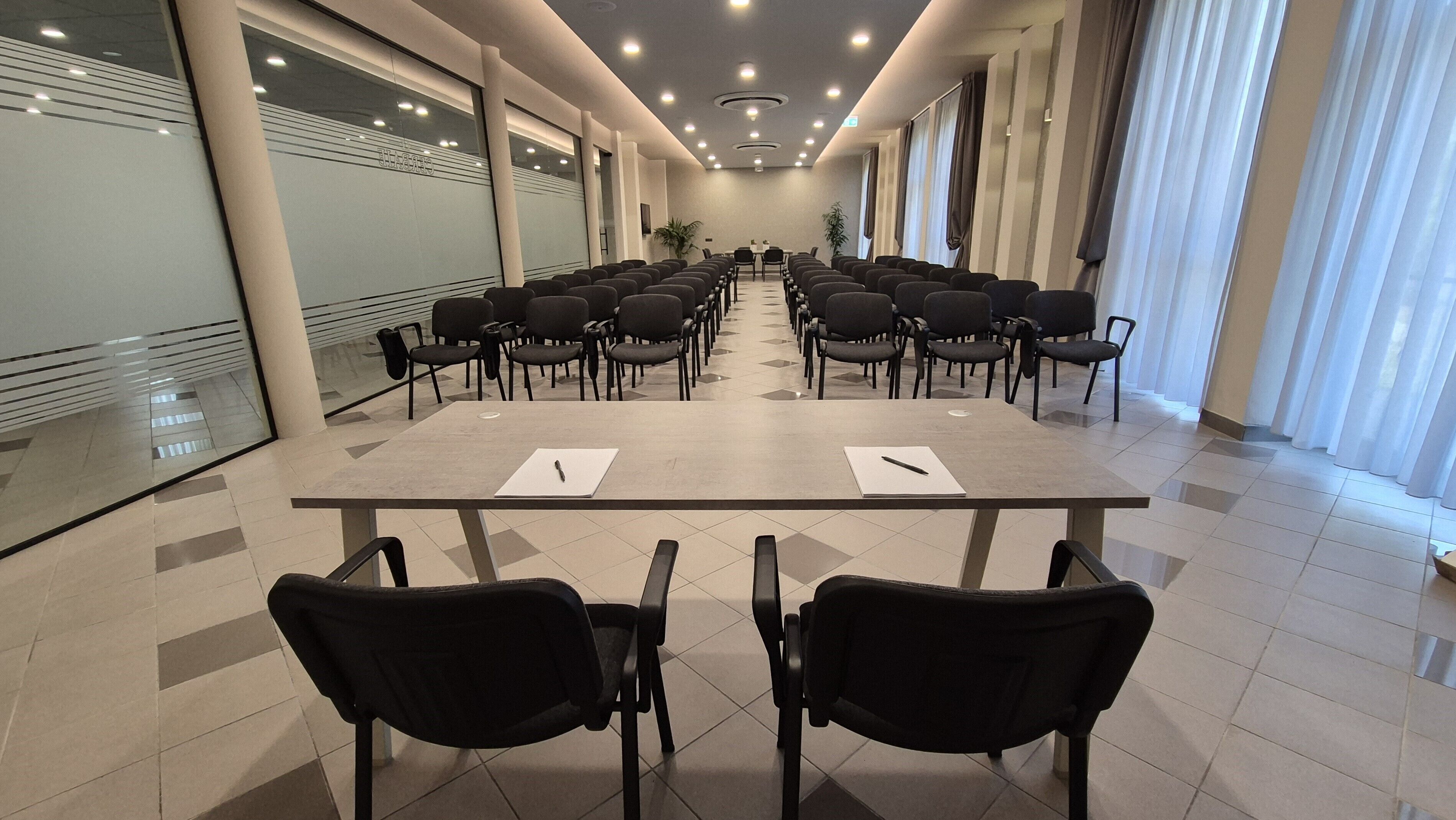 Un salón de conferencias con una larga mesa y sillas, ventanas amplias con cortinas y suelo de baldosas.