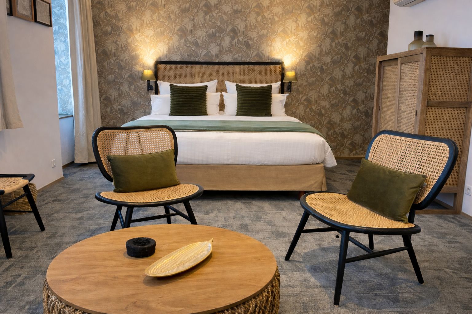 Un dormitorio con una cama, dos sillones de mimbre, una mesa redonda de madera y una pared con estampado floral.