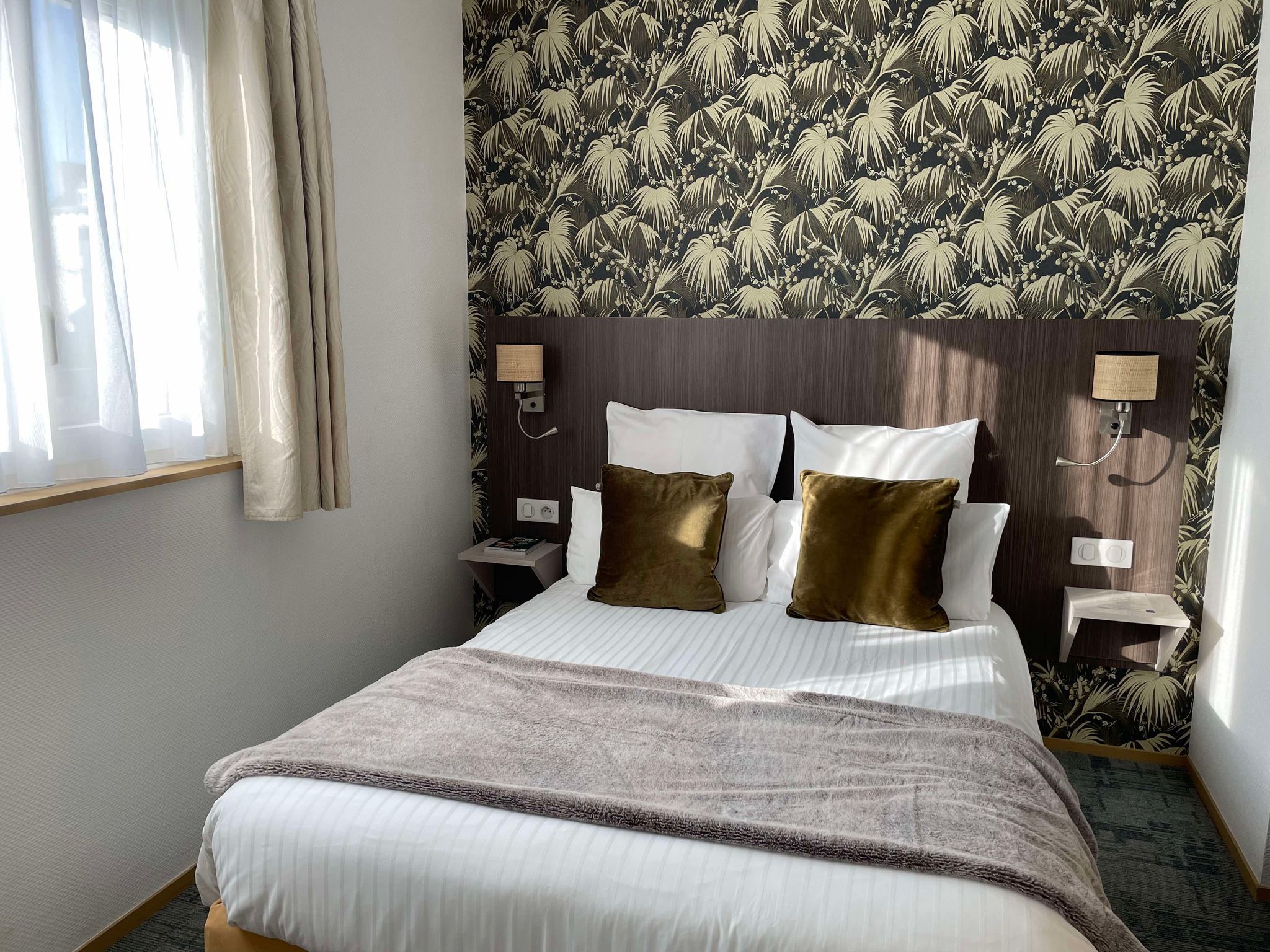 A hotel room with a large bed, two bedside tables, a nightstand, a wall-mounted lamp, and a window with curtains.