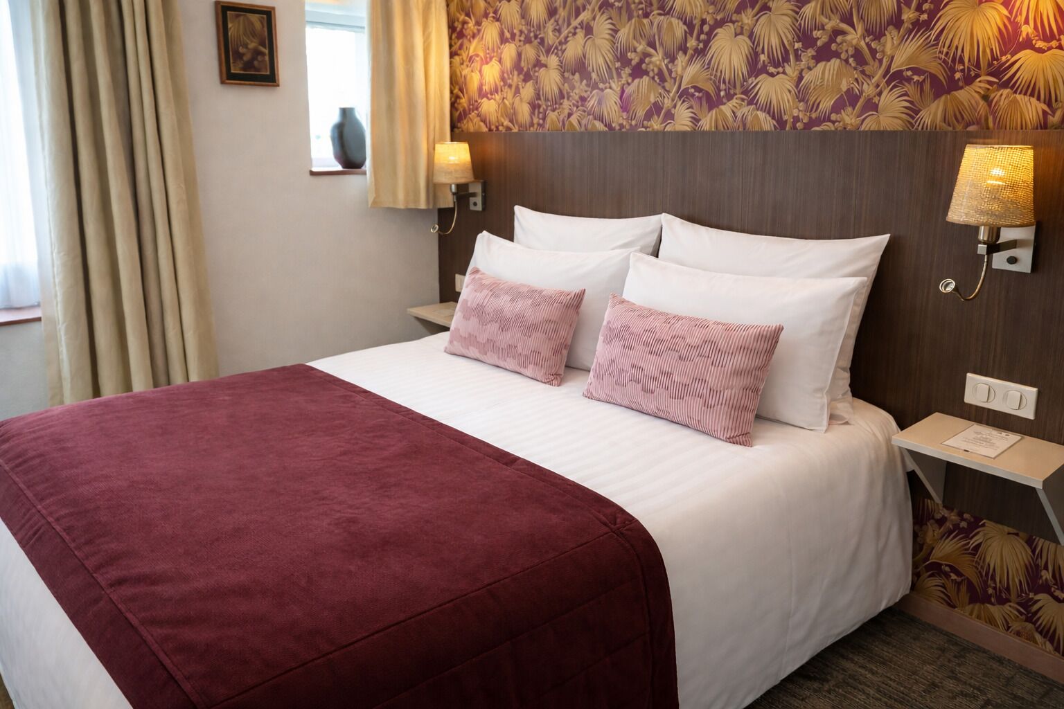 Habitación de hotel con cama, dos almohadas, mesita de noche, lámpara y un papel tapiz con diseño floral.