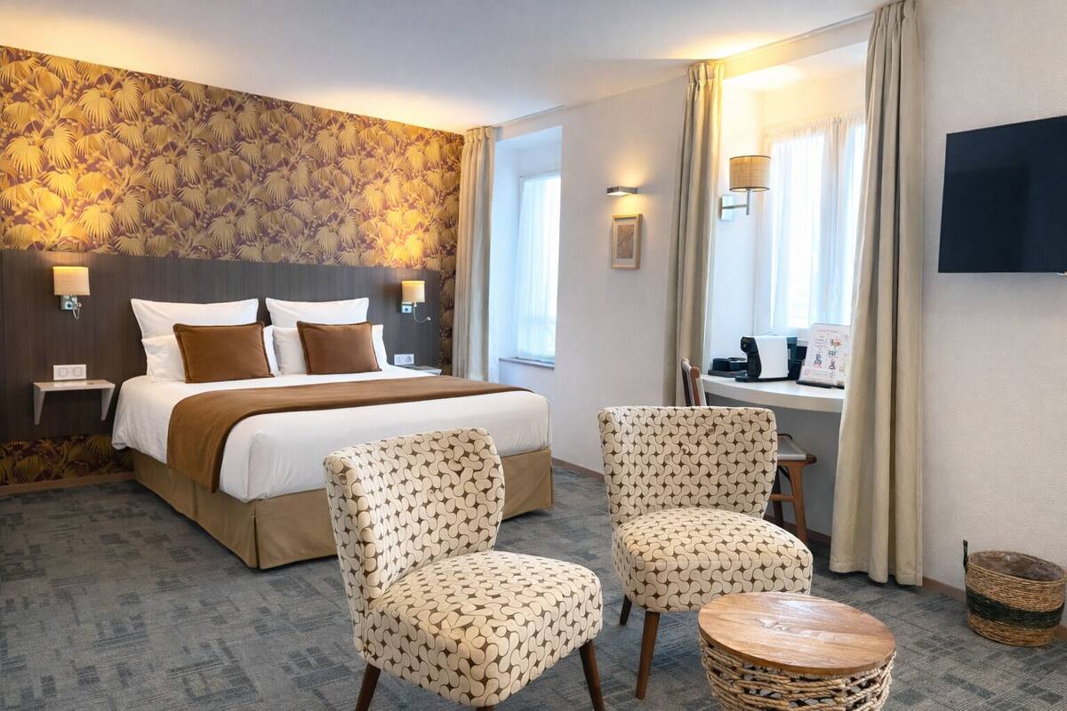 Una habitación de hotel con una cama, dos sillones estampados, una mesita, un televisor y un ventanal con cortinas.