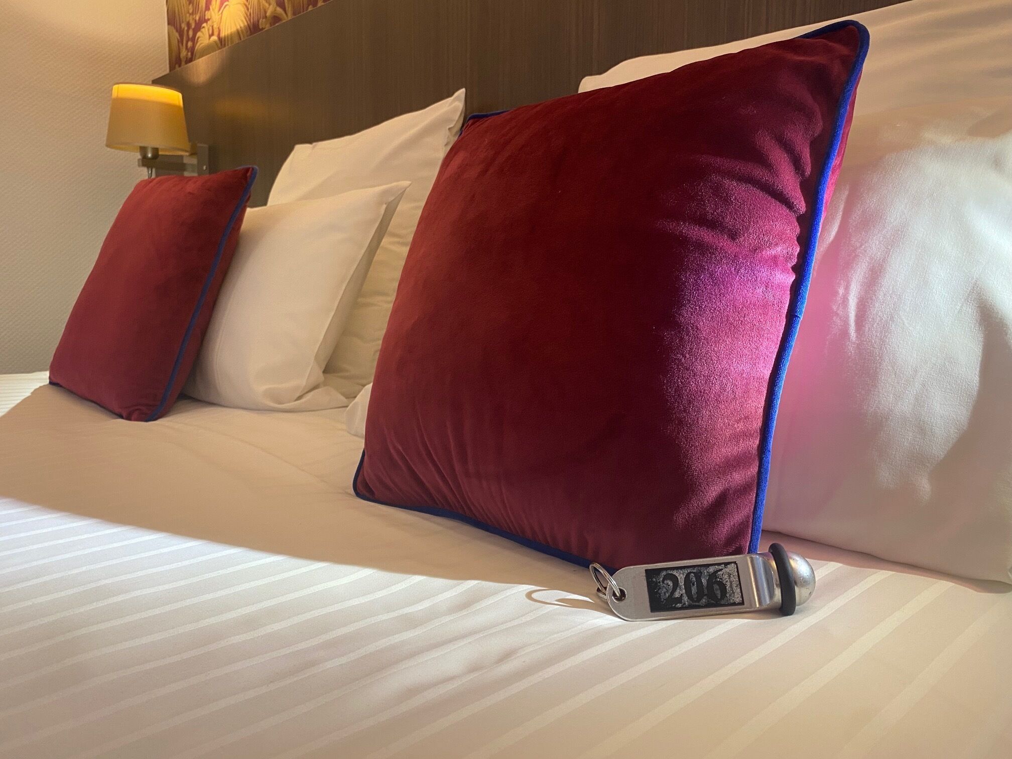 Una cama de hotel con sábanas blancas, almohadas rojas y un reloj digital que marca las 20:06.