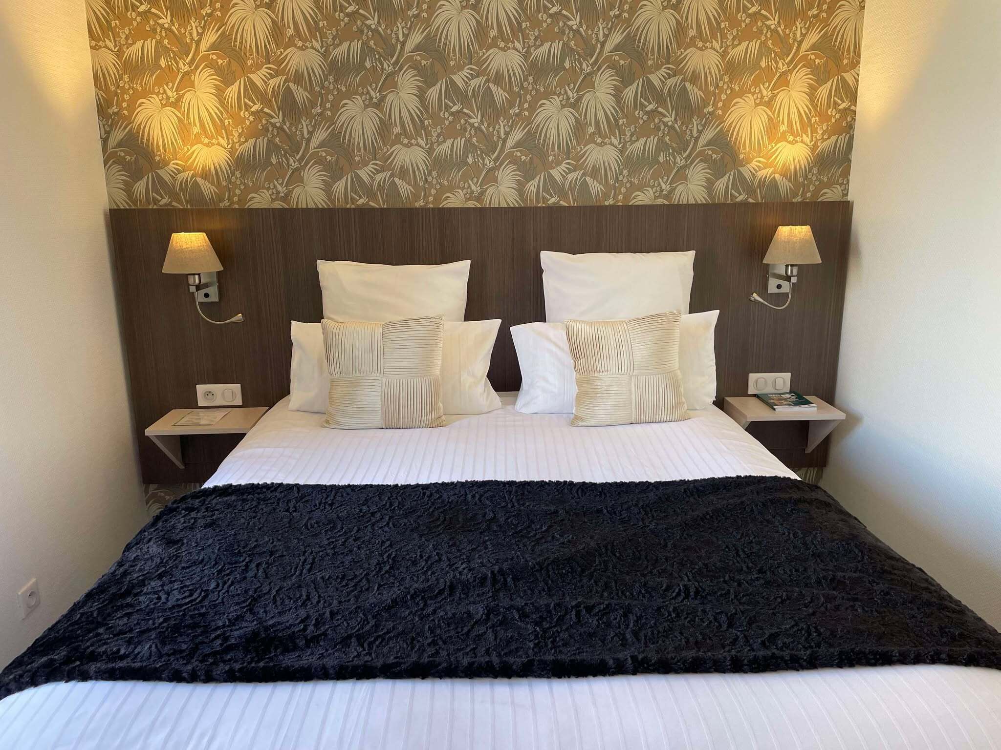 Quarto de hotel com uma cama grande, duas luminárias de cabeceira, uma mesa de apoio e uma parede com padrão de folhas.