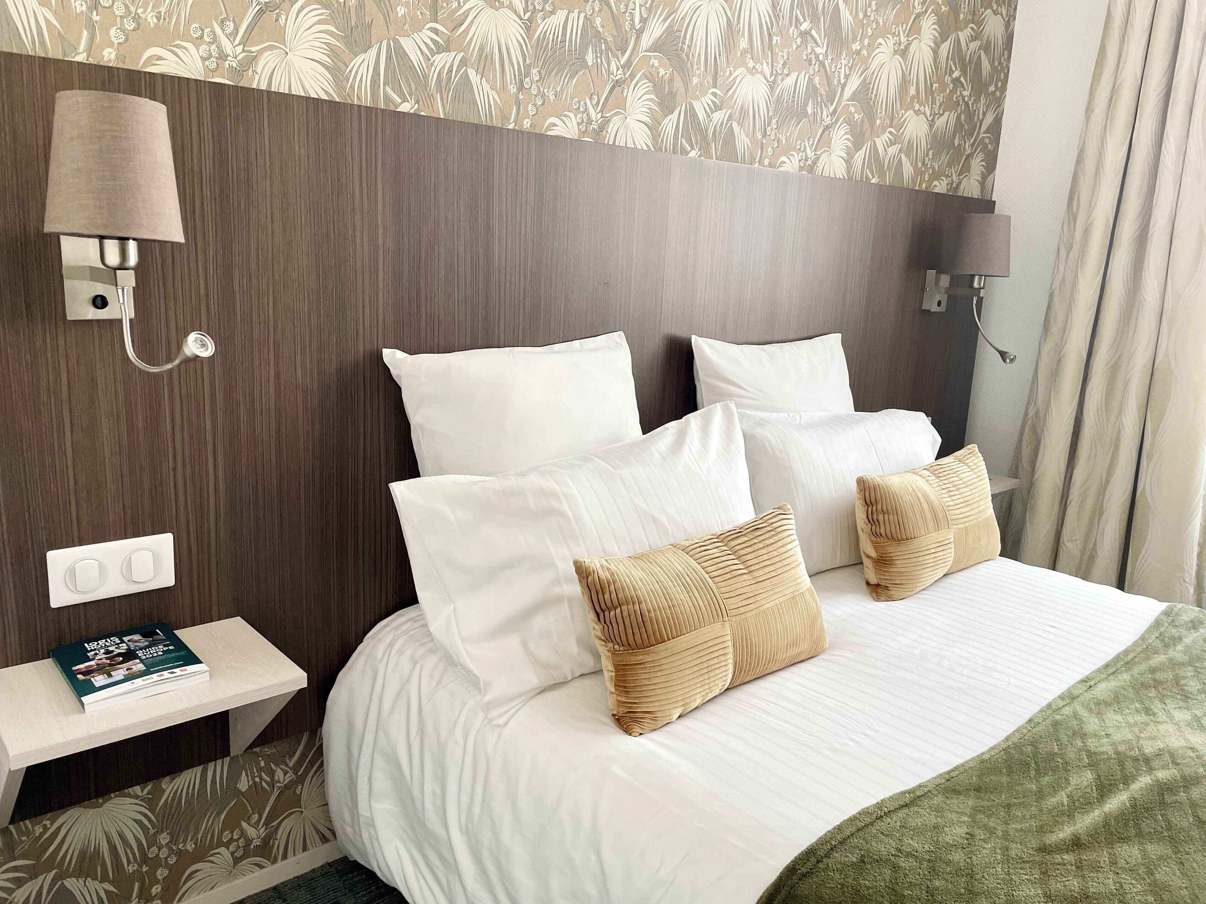 Hotellihuone, jossa on puinen sängynpääty, valkoiset vuodevaatteet ja pieni yöpöytä, jonka päällä on kirja.