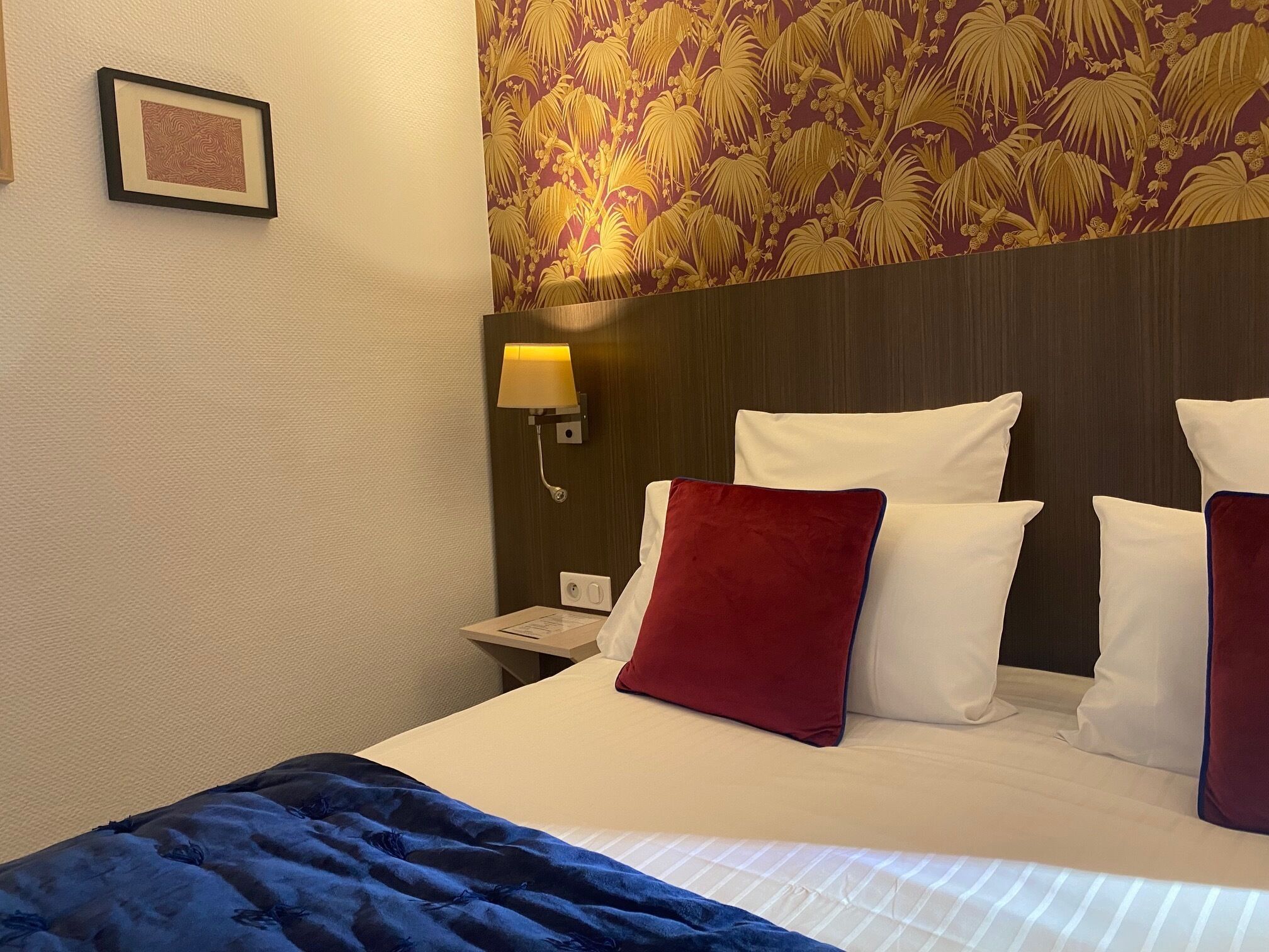 A hotel room with a bed, white pillows, a red pillow, a blue blanket, a nightstand with a lamp, and a floral wallpaper.