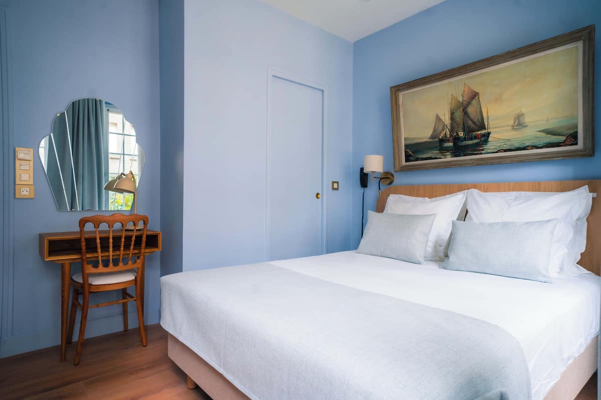 A bedroom with a bed, a desk, a chair, a mirror, and a painting on the wall.