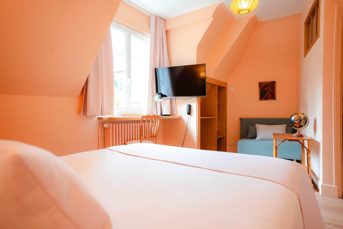 A hotel room with a large bed, a television mounted on the wall, a window with curtains, a radiator, and a small table with a globe.